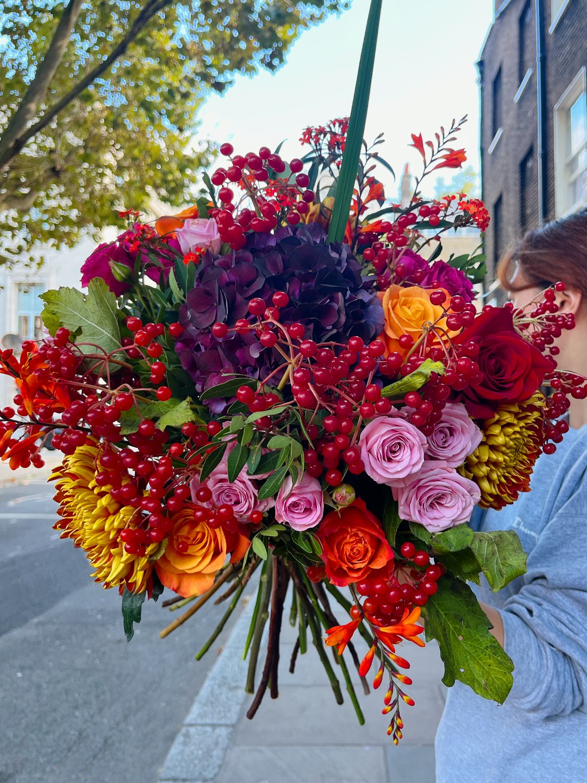A vibrant bouquet of autumn flowers including roses, chrysanthemums, deep hydrangeas, and bold red berries, wrapped in a complimentary bouquet bag with water.
