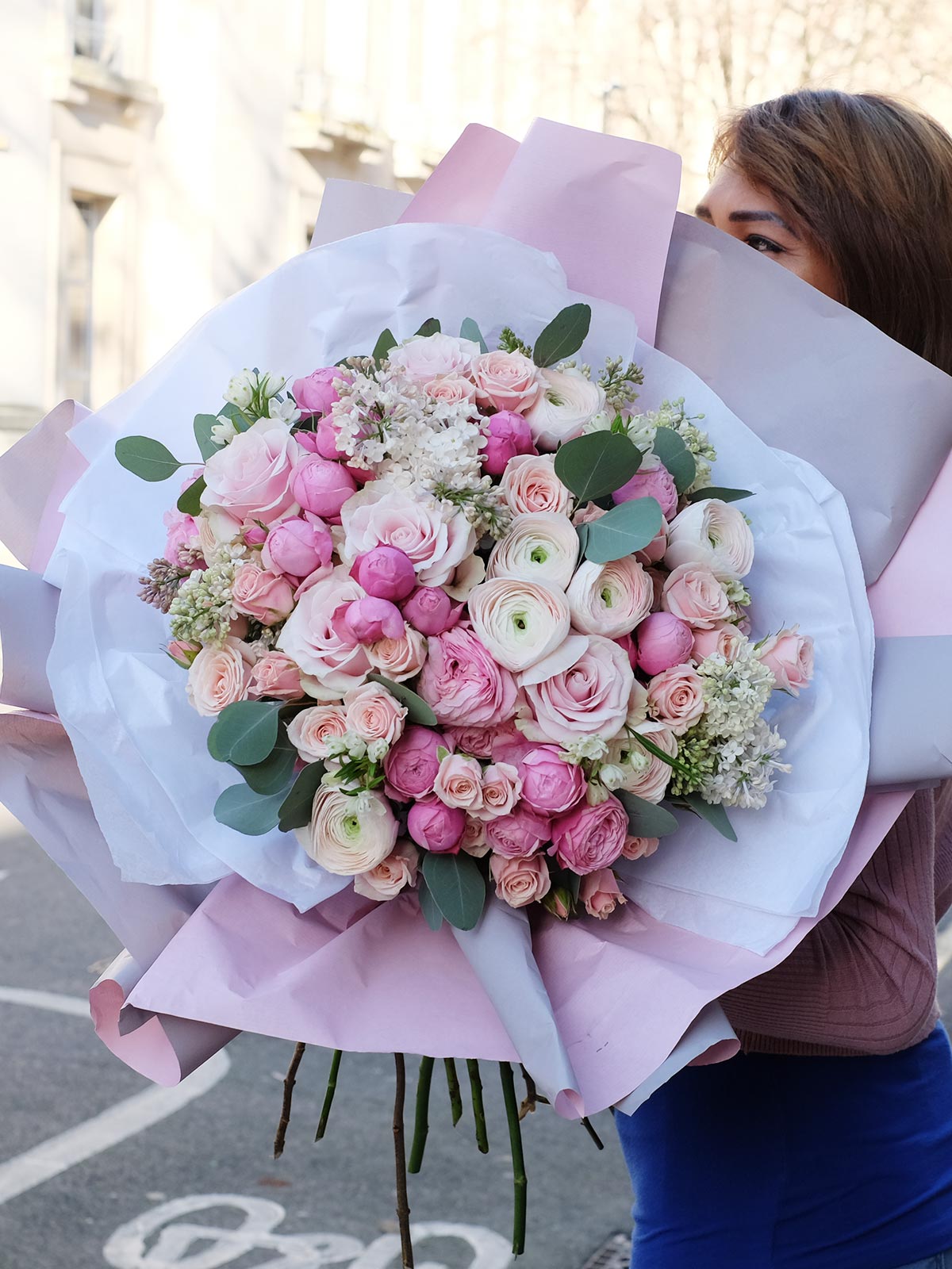 A person holding a large bouquet of flowers primarily in shades of pink, with greenery, wrapped in pink and white paper.