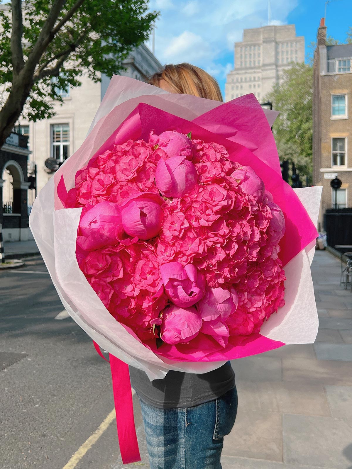 A person holding a large bouquet of juicy pink peonies and lush hydrangeas, wrapped in pink and white paper.