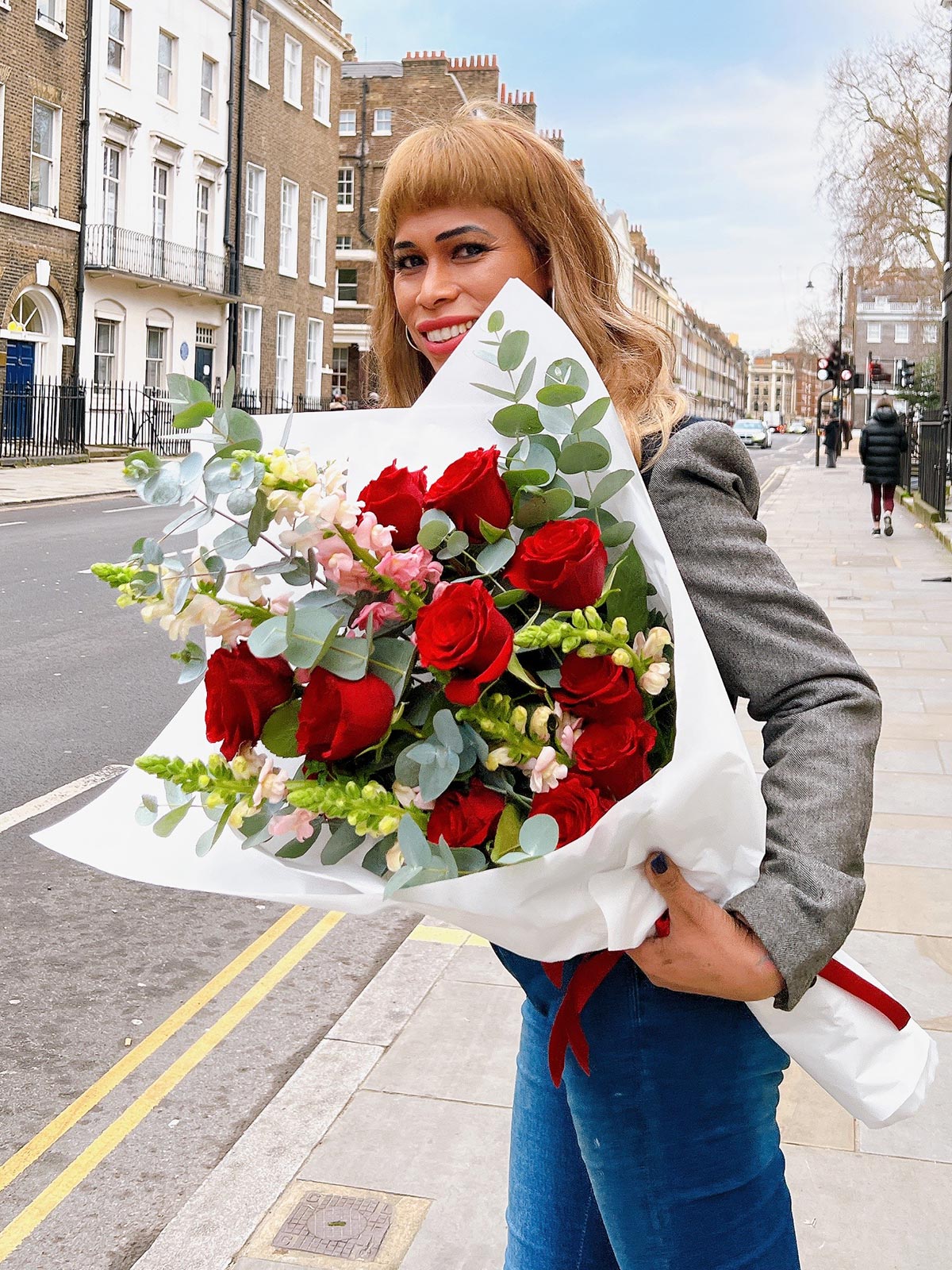A person holding a classic sized bouquet of deep red roses, eye-catching dragonsnaps, and fresh eucalyptus with a white wrap and a red ribbon.