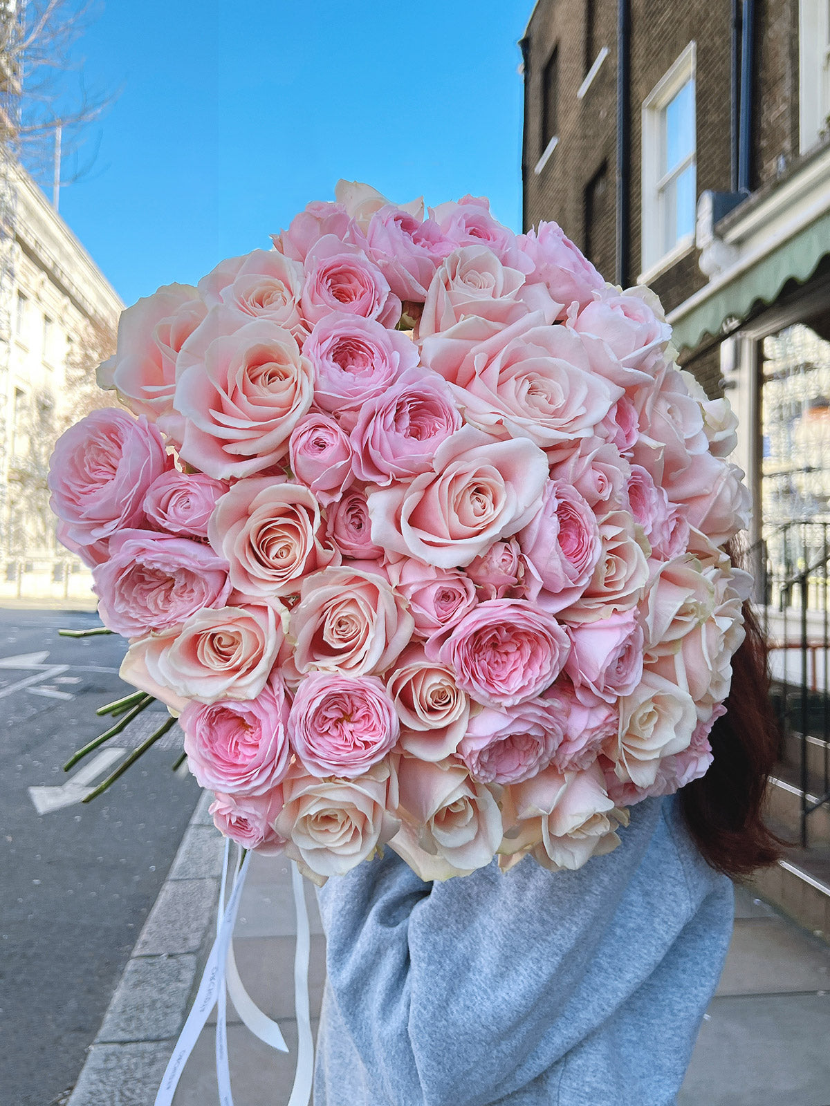 A bouquet of pink roses and peony roses, predominantly in shades of pink, wrapped with a light blue paper ribbon.